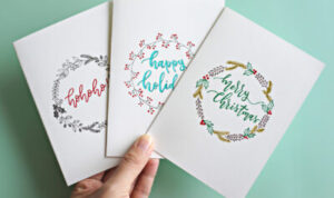 Cropped Hand Holding Christmas Cards With Text Over Turquoise Background